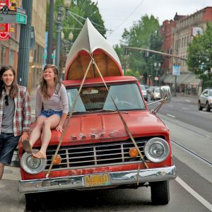 two people next to an orange truck with a canoe strapped to the top