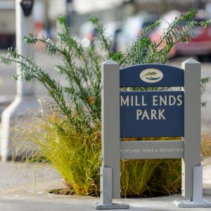 A wooden sign reading \"Mill Ends Park Portland Parks & Recreation\" stands on a concrete ring enclosing a small planter filled with an evergreen bush and light green grass