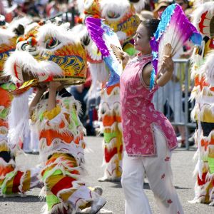 women dances with large fans outside on a city street surrounded by people in colorful dragon costumes