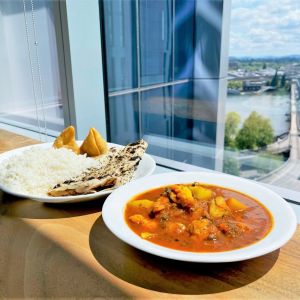 A white plate of aloo gobi sits next to a platter of rice, samosas and naan on a wooden table in front of a large window.