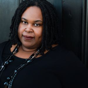 A Black woman with dark mid-length twists, a beaded necklace and a black shirt subtly smiles into the camera.