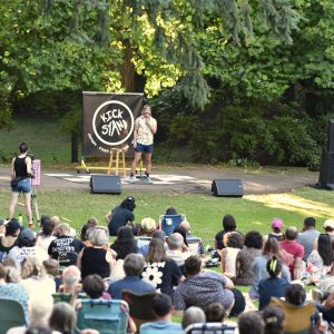 Blond comedian on stage performing in front of large crowd sitting in a park