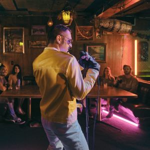 a person in a yellow leather-looking jacket singing in to a microphone in front of patrons in a bar