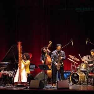 A band of five Jazz musicians performs on stage in front of a red curtain.