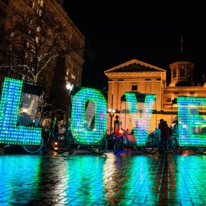 a night image of the word LOVE spelled out in large letters made of hundreds of small bulbs attached to bicycles in front of a large brick building