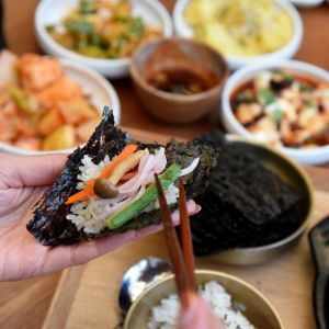 chopsticks reach for colorful vegetables atop a sheet of seaweed with a spread of Korean food in the background