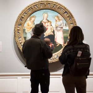 two people and a baby stand in front of a round oil classical painting of a religious scene at the Portland Art Museum