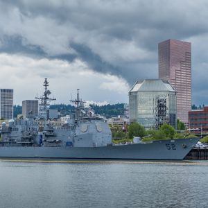 large warship docked at Waterfront Park in Portland with dramatic grey clouds over buildings in downtown