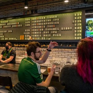 people sitting at a bar, a large board behind lists beers on draft