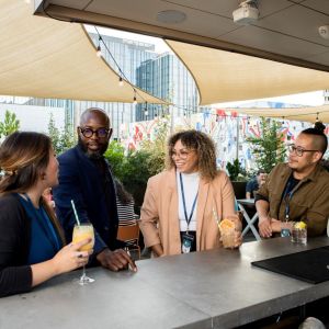 Four people stand at a bar talking, three have drinks.
