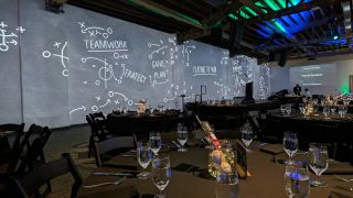 an event stage with large projection wall showing chalkboard diagrams with words including TEAMWORK and STRATEGY behind it