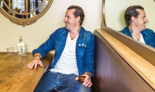 A man is sitting in a stylish restaurant booth, holding an espresso and smiling.