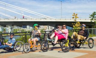 Four people ride three-wheeled bicycles along a river; two of the bikes are hand-operated