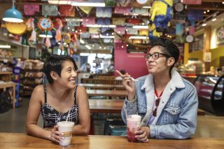 Two smiling people sit at a wooden table in a brightly decorated interior