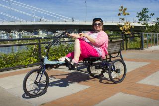 A woman pedals a recumbent three-wheeled bike with a bridge in the background