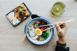 a hand holding chopsticks hovers over a bowl of ramen. a mug of green tea and a plate of fried chicken are next to the ramen bowl on a table.