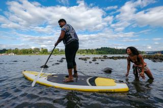 A woman pilots a stand-up paddleboard in a river, with another woman looking on