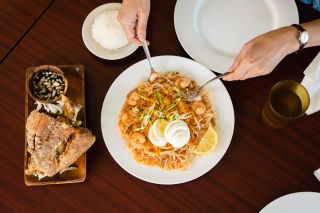 Two hands serve up a shrimp noodle dish, next to a crispy piece of pork