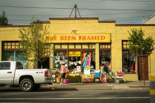 Two people shop sidewalk offerings in front of a bright yellow storefront with a sign reading "I've been framed"