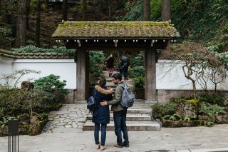 A woman and man wearing backpacks and winter coats stand before a stone gate surrounded by greenery