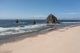 the Oregon coast on a sunny day, with waves rolling toward a sandy beach, around huge rock formations, beneath a blue sky