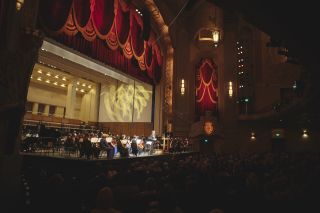 side view of symphony players on the stage at a large hall with dimmed house lights