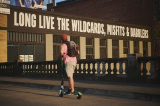 person on e-scooter passes mural reading Long Live the Wildcards, Misfits & Dabblers