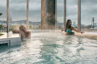 Two women sitting in a hot tub with a view of bridges over the Willamette in the background.