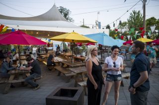 people stand in a food cart pod patio with colorful umbrellas and flags covering picnic tables