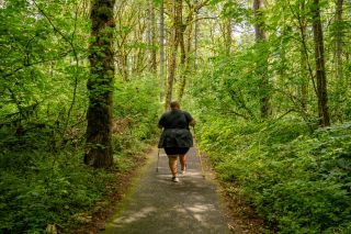 Wide back shot of a disabled Black non-binary hiker entering a forest with trekking poles via an accessible paved trail. They are surrounded by lush greenery, with sunlight peeking through the back of the visible path.