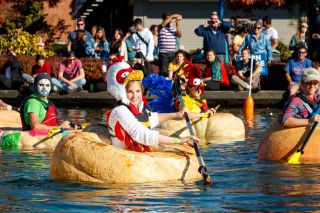 people in costumes paddle boats made of giant pumpkins on a lake while spectators watch on the shore
