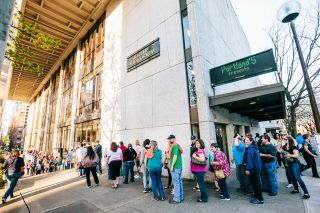 people stand in a line on a city sidewalk as they wait for entry into the Keller Auditorium