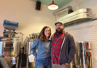 A couple stands in front of craft brewing barrels inside a small room lined with subway tiles.