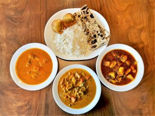 Three dishes of curries and aloo gobi sit next to a platter of rice, samosas and naan on a wood table.