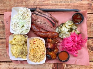 a tray of barbecued meat, sauces, sides of pickle and cucumber, and coleslaw, mashed potatoes and macaroni and cheese