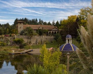 a large stone building overlooks a Portland vineyard and a pond, with trees all around and wispy clouds above