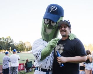 A giant pickle wearing a baseball uniform and hat poses for a photo with a man wearing a Portland Trailblazers shirt