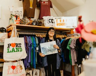 a woman surrounded by Portland tote bags and other Portland clothes and merch holds up a print of a Portland bridge