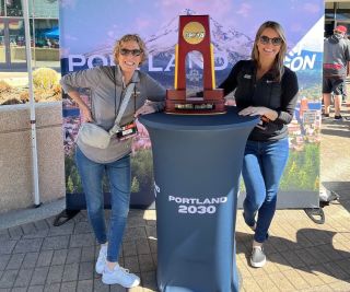 two women pose with the NCAA Women's Final Four trophy sitting on a podium that says Portland 2030
