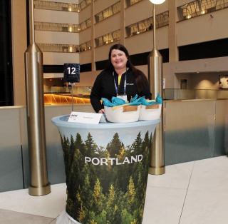 A woman stands at a pedestal table adorned with a forest image and the word PORTLAND.