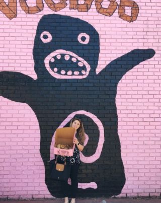 photo of person with donuts in front of pink wall with Voodoo Donut sign