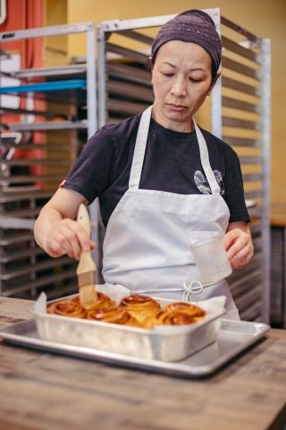 a person wearing a head covering and an apron prepares a tray full of large cinnamon rolls