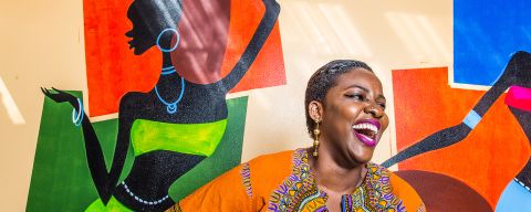 a Black woman wearing an orange dashiki stands in front of a colorful mural laughing