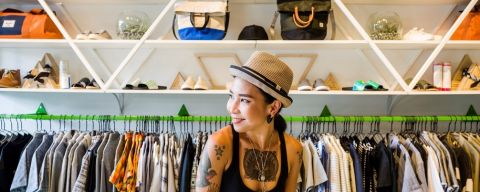 a woman with tattoos and wearing a hat stands in front of a rack of clothing at Wildfang clothing store