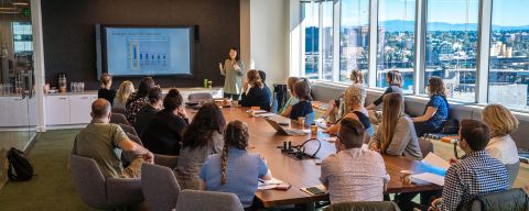 a large group of people around a conference room table watch a person giving a presentation