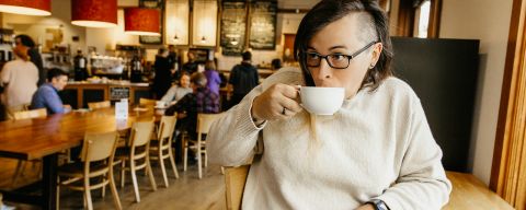 a white person with long hair on one side and a shaved head on the other side, sips from a coffee mug in a warmly lit bakery.