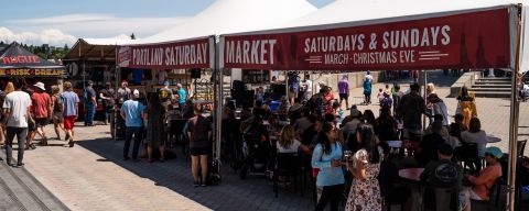 People stand under a canopy with signs reading \"Portland Saturday Market Saturdays & Sundays March - Christmas Eve\"