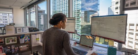 a woman works at a computer with two large monitors in front of a window with a city skyline view