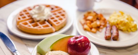 table with traditional breakfast foods, waffle, home fries, sausages, scrambled eggs and fruit with a pastry