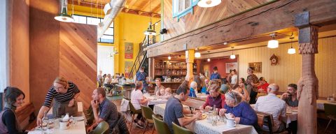 tables full of diners enjoy a meal in a lively, warmly lit restaurant with a bar in the background